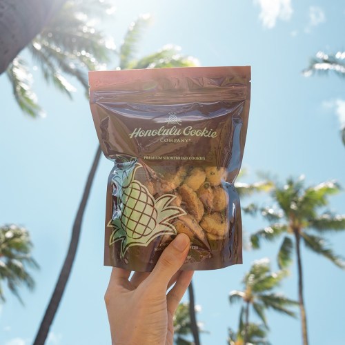 A hand holds a bag of Honolulu Cookie Company shortbread cookies against a sunny sky and palm trees.