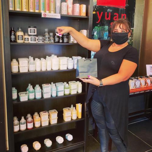 A woman in a black mask holds a gift bag while reaching for a product on a shelf filled with various skincare items.