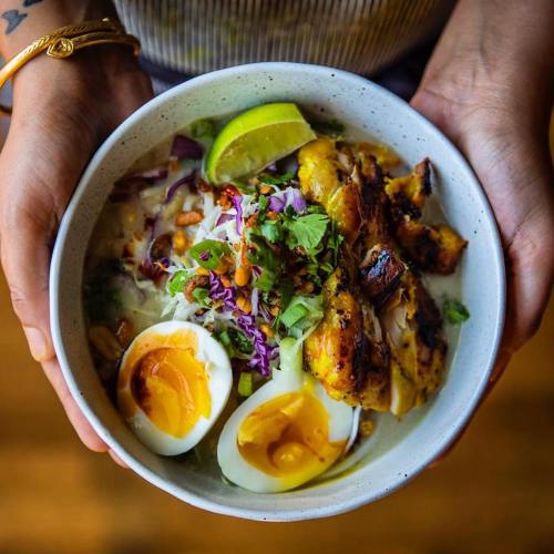 A person holds a bowl of soup topped with grilled chicken, soft-boiled eggs, herbs, and lime.