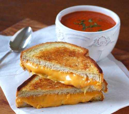 A grilled cheese sandwich with melted cheese, served alongside a bowl of tomato soup garnished with herbs.