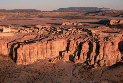 Aerial view of a desert landscape featuring a cliffside village surrounded by rocky terrain and distant mountains.