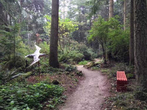 A winding forest path surrounded by lush greenery, with a red bench and a bird sculpture nearby.