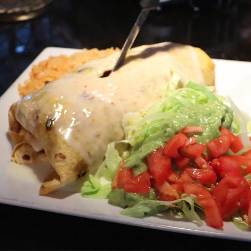 A plate featuring a large enchilada topped with cheese sauce, served with rice, lettuce, tomatoes, and guacamole.