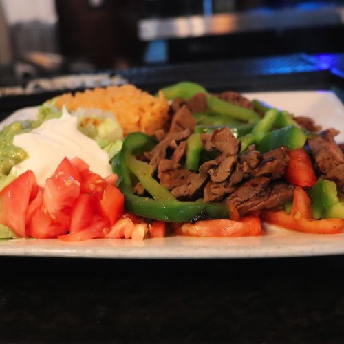A plate of beef fajitas with green peppers, tomatoes, lettuce, guacamole, sour cream, and rice.