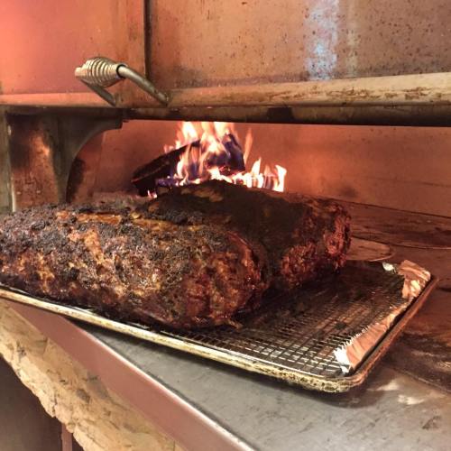 A large, smoked meat brisket cooking in a barbecue pit with flames visible in the background.