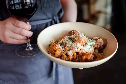 A person holds a bowl of spaghetti and meatballs topped with cheese, alongside a glass of red wine.