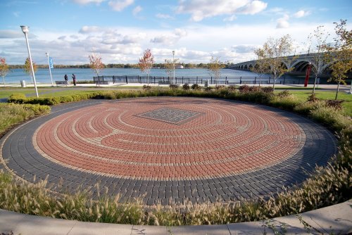 A circular brick labyrinth surrounded by greenery, with a waterfront and bridge in the background under a cloudy sky.