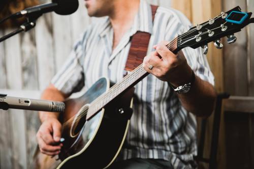 A musician plays an acoustic guitar while singing into a microphone, seated against a wooden backdrop.