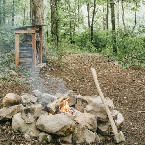 A campfire surrounded by stones, with an axe nearby, set in a lush green forest. A wooden structure is in the background.
