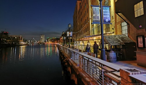A waterfront scene at dusk with lights reflecting on the water, featuring signs for beer and food along the boardwalk.