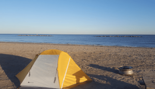 A yellow tent on a sandy beach with calm water and a rocky jetty in the background under a clear blue sky.