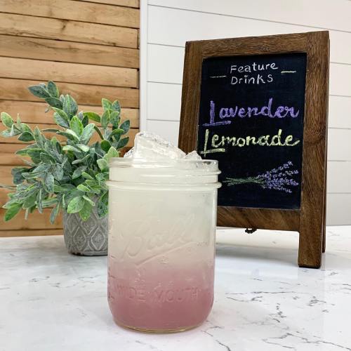 A glass of lavender lemonade with ice, next to a chalkboard sign displaying the drink's name and a plant in the background.