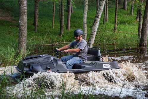 A person in a helmet rides a black hovercraft through a flooded area, creating splashes in the water.