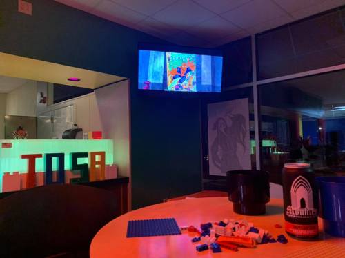 A cozy room with colorful lighting, a TV displaying art, a table with LEGO pieces, and a can of Monks beer.