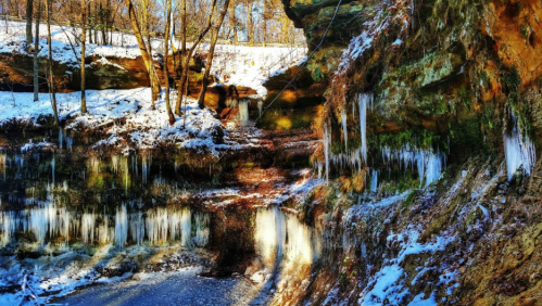 A snowy landscape with icicles hanging from rocky cliffs, surrounded by trees in a serene, winter setting.