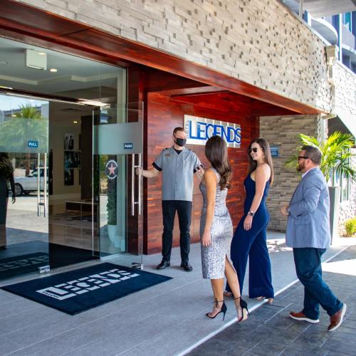 Two women in elegant outfits and a man in a suit enter a building labeled "LEGENDS," greeted by a staff member.