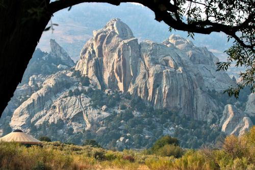 A rugged mountain landscape with large rock formations, framed by trees and a yurt in the foreground.