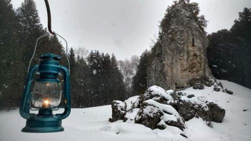 A blue lantern hangs in the foreground, with a snowy landscape and a rocky cliff in the background. Snowflakes are falling.