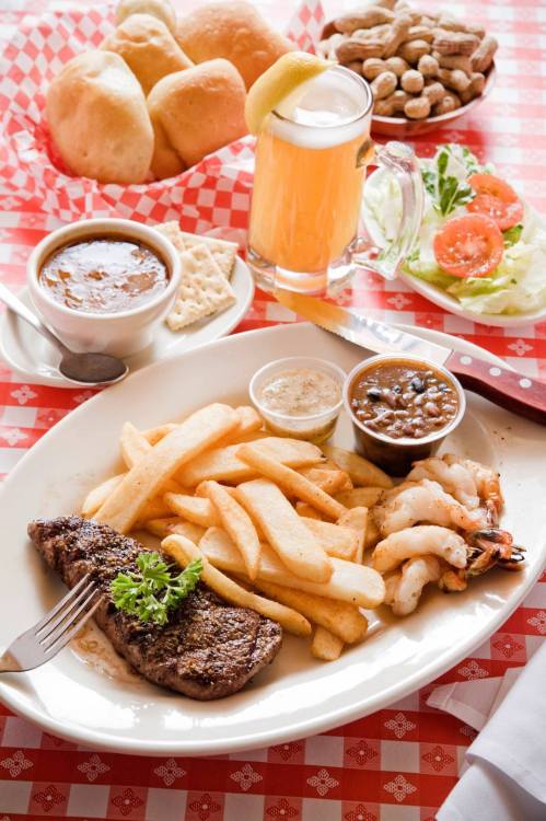 A plate with steak, shrimp, fries, and sauces, accompanied by a bowl of soup, salad, bread rolls, and a drink.