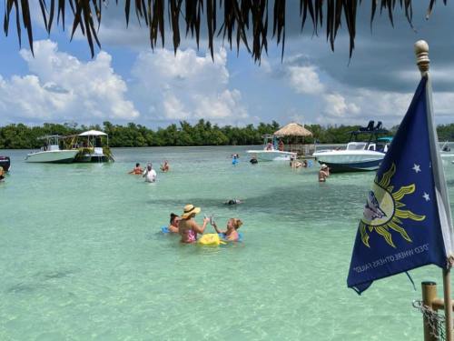 People enjoying a sunny day in shallow water, with boats nearby and a flag waving in the foreground.