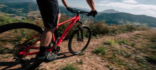 A cyclist rides a red mountain bike on a dirt trail surrounded by hills and greenery under a blue sky.