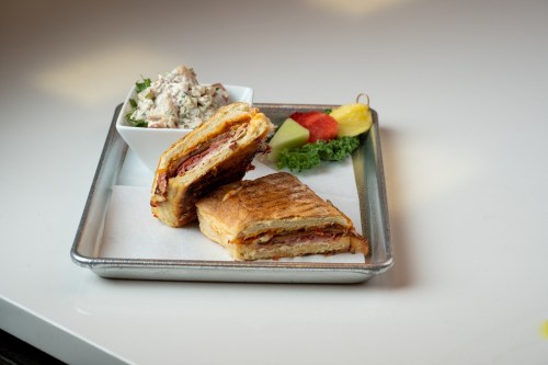 A toasted sandwich cut in half, served with a side of salad and assorted fruit on a metal tray.