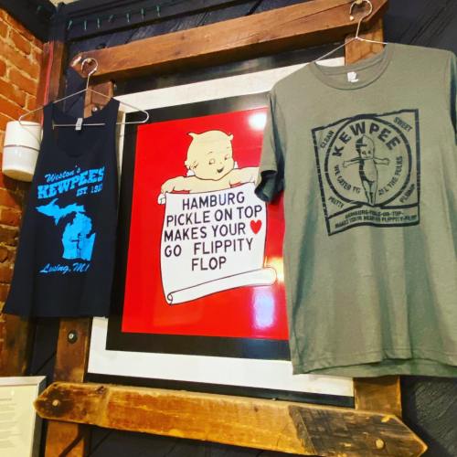 Two T-shirts hang beside a colorful sign that reads, "Hamburg pickle on top makes your heart go flippity flop."