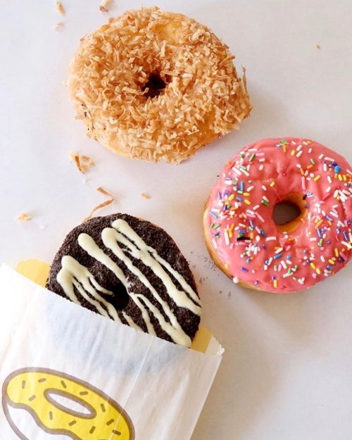 Three colorful donuts: one with pink icing and sprinkles, one chocolate with white drizzle, and a coconut-covered donut.
