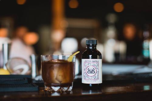 A cocktail glass with a lemon twist next to a bottle of bitters on a bar counter, with a blurred background.