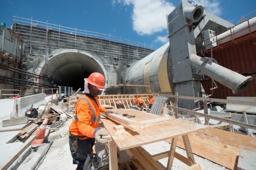 A construction worker in an orange vest and hard hat works on a wooden structure near a tunnel under construction.