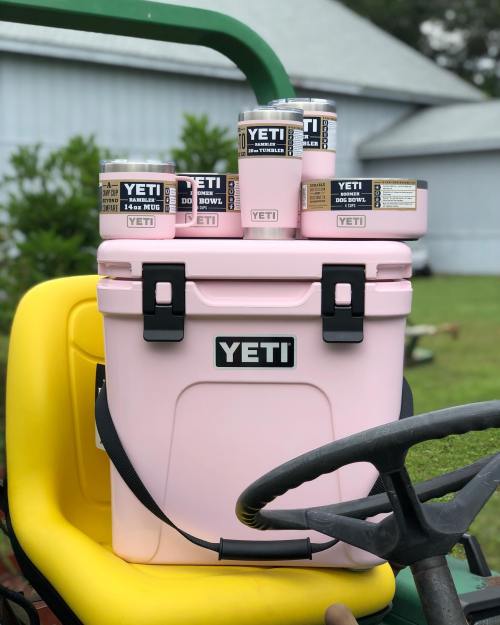 A pink YETI cooler with matching drinkware sits on a yellow tractor seat in a grassy outdoor setting.