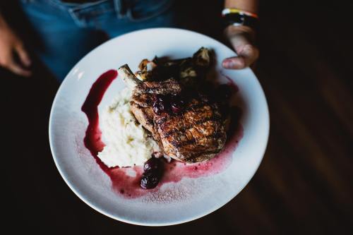A person holds a plate with grilled meat, mashed potatoes, and a berry sauce, set against a dark background.