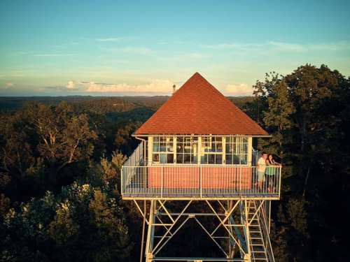 A scenic view of a lookout tower surrounded by trees, with two people standing on the balcony at sunset.