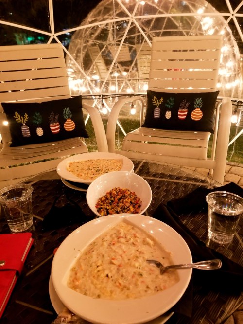 A cozy outdoor dining setup with two bowls of soup, a salad, and decorative pillows, illuminated by string lights.
