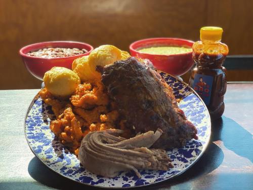 A plate of BBQ meat with sides of cornbread, beans, and a bottle of sauce, set against a wooden background.
