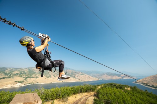 A person zip-lining over a scenic landscape with mountains and a lake under a clear blue sky.