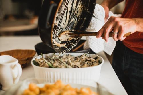 A person stirs a creamy dish in a white baking dish, with a skillet and another dish in the background.