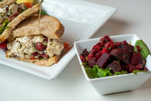 A sandwich with chicken salad and cranberries, served with a side of beet and herb salad in a white bowl.