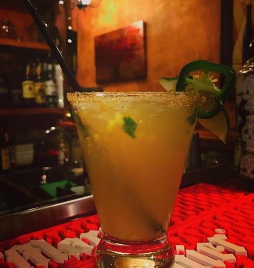 A refreshing cocktail garnished with jalapeño and lime, served in a glass with a salted rim on a bar counter.