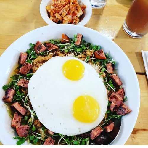 A bowl of noodles topped with two sunny-side-up eggs, diced meat, sprouts, and a side of kimchi.