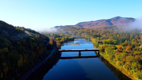 Aerial view of a river surrounded by colorful autumn foliage and mountains under a clear blue sky.