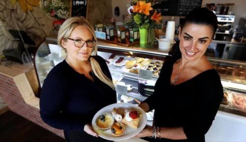 Two women hold a plate of desserts in a café, with a display case of pastries and flowers in the background.