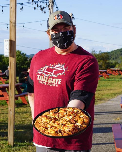 A person wearing a mask and a maroon shirt holds a pizza on a tray outdoors, with picnic tables in the background.
