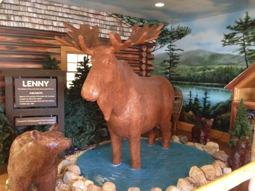 A large moose statue named Lenny stands in a pond, surrounded by trees and a cabin, with two bear statues nearby.