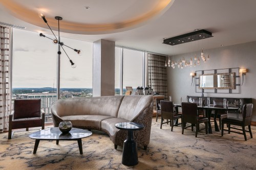 A modern hotel suite featuring a curved sofa, dining area, and large windows with a city view. Elegant decor throughout.