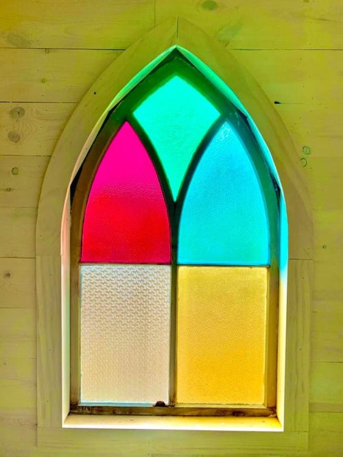 Colorful stained glass window with a gothic arch design, featuring pink, green, blue, and yellow sections.