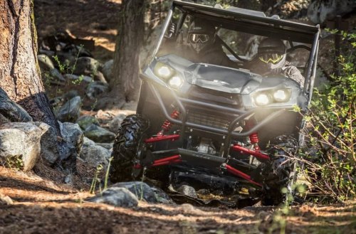 A black ATV navigating a rocky forest trail, with two riders wearing helmets and goggles. Dust is kicked up behind it.