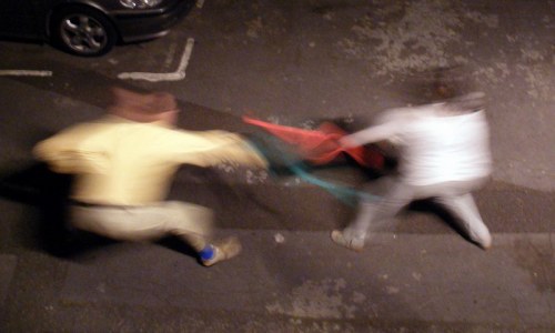 Two blurred figures appear to be playfully tugging on a red object in a dimly lit street.