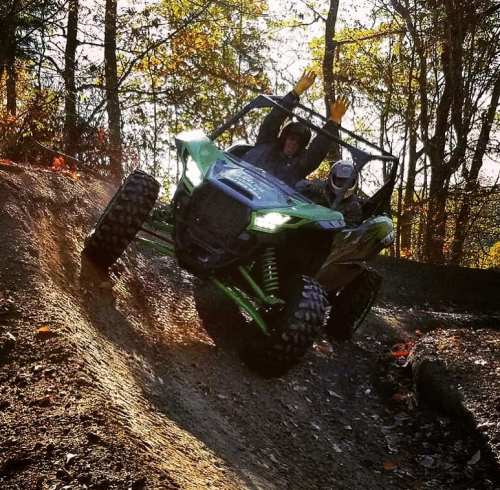 Two people ride an ATV on a dirt trail, raising their arms in excitement amidst autumn trees.