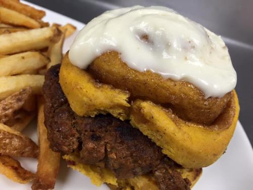 A burger topped with a cinnamon roll and cream cheese frosting, served with a side of French fries.